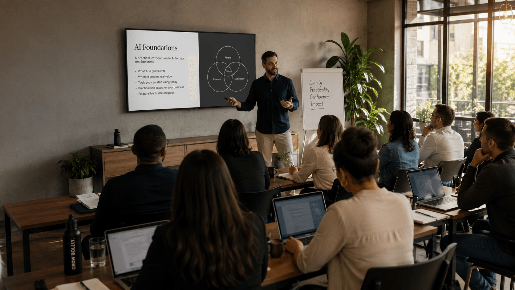 A trainer presenting an AI Foundations slide to a seated group taking notes on laptops in a bright workshop space.
