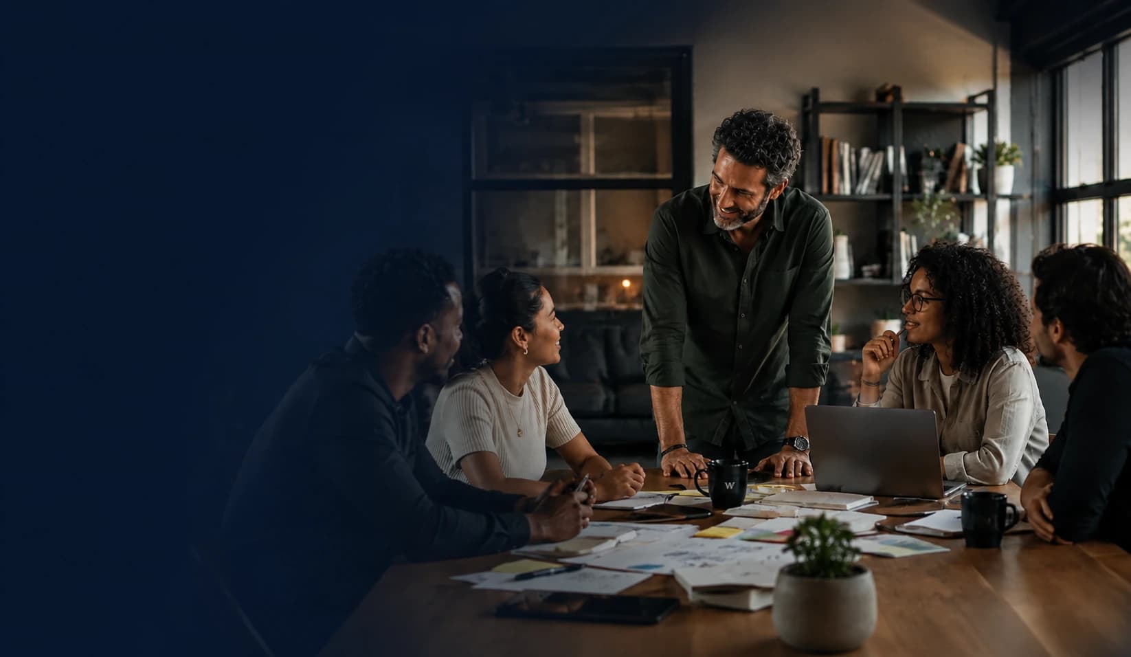 A team of five colleagues collaborating around a table during a strategy discussion, with a leader standing and gesturing as colleagues review notes and a laptop, set against calm negative space to the left.
